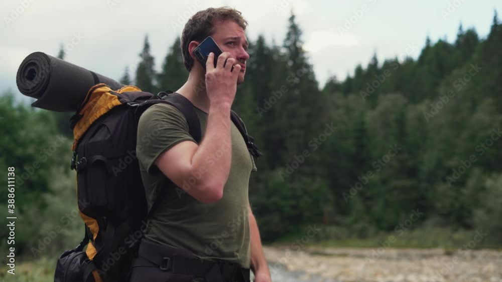 a man tourist with a backpack behind his shoulders stands near the river dials a number on a smartphone and makes a call a man tourist with a backpack behind his shoulders stands near the river dials a number on a smartphone and makes a call