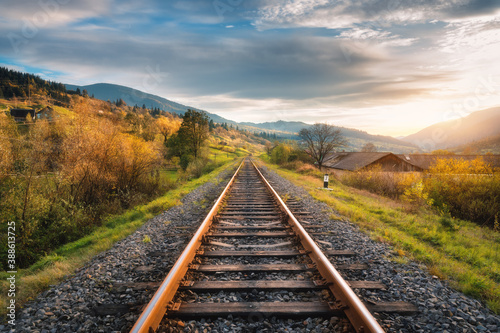 Railroad in mountains at sunset in autumn. Beautiful industrial landscape with railway station, orange trees, green grass, buildings, rocks and blue sky with clouds in fall. Rural railway platform