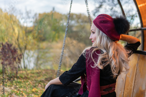 A beautiful woman sits on a garden swing, forged in a burgundy coat and biret, an adult smiles at the camera, in the fall against a background of trees, a blue sky.