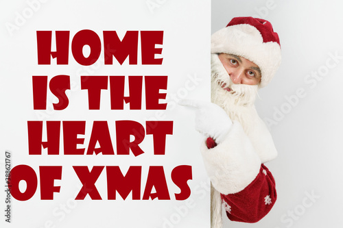 Santa Claus points his fingers at the board with the text - HOME IS THE HEART OF XMAS