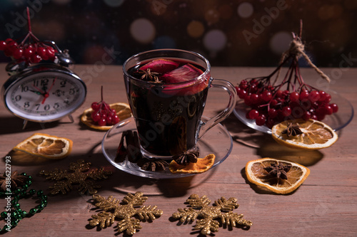 A Cup of fragrant mulled wine with spices, decorations for the New year, a branch of berries on a plate and a clock showing twelve o'clock. Horizontal orientation