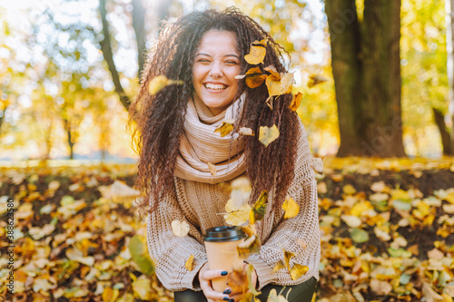Positive afro hair woman with beautiful smile wearing knitted sweater and scarf throw yellow autumn leaves in park on sunny day.
