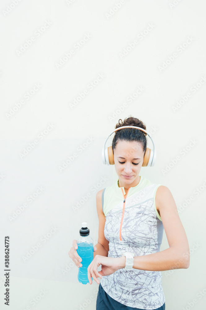 Naklejka premium young female athlete looks at watch while carrying energy drink and listening to music with headphones
