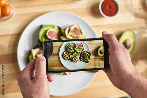 Close-up of man holding mobile phone and making photo of sandwiches on the plate with fresh vegetables