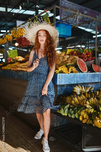 Beautiful young girl in a straw hat and a blue dress stands near boxes of bananas in an asian market. Thailand, Phuket © Nikolay