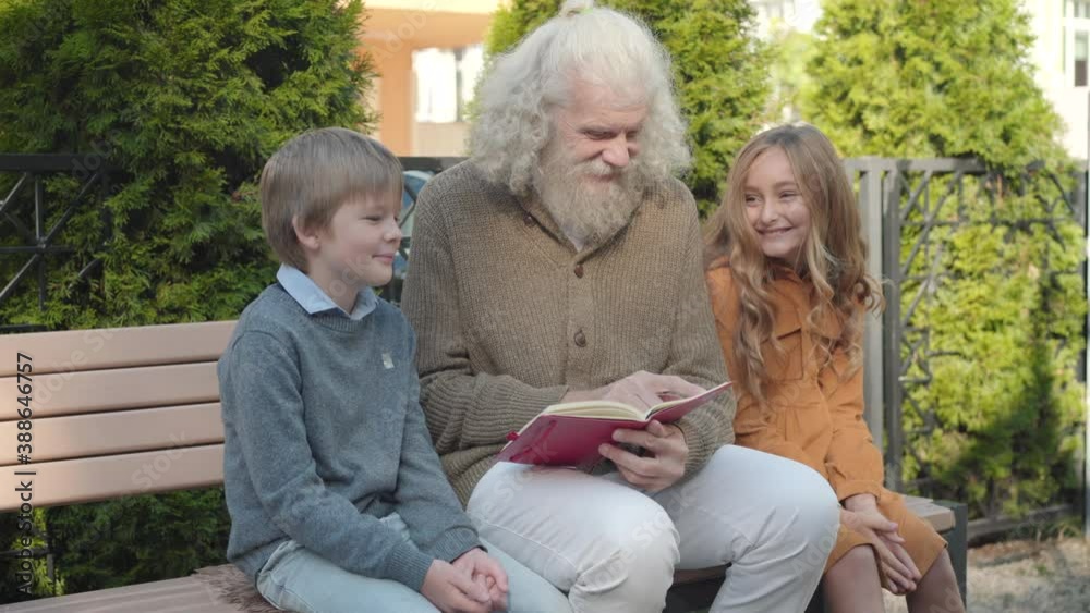Positive grandfather sitting with grandchildren on bench and reading book. Portrait of happy relaxed Caucasian old man, little girl and boy enjoying sunny weekend outdoors.