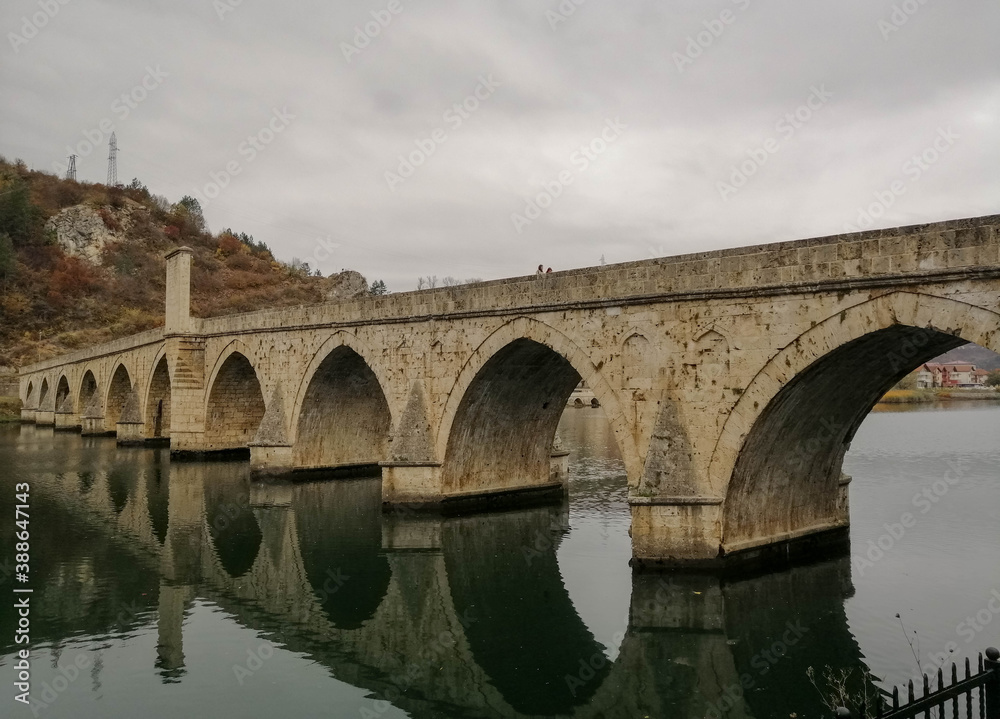 Beautiful old stone bridge with arches diagonnally over a river reflecting in the water, autumn scene