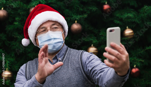 Christmas online greetings. Close up Portrait of senior man wearing a santa claus hat and medical mask with emotion. Against the background of a Christmas tree. Coronavirus pandem photo