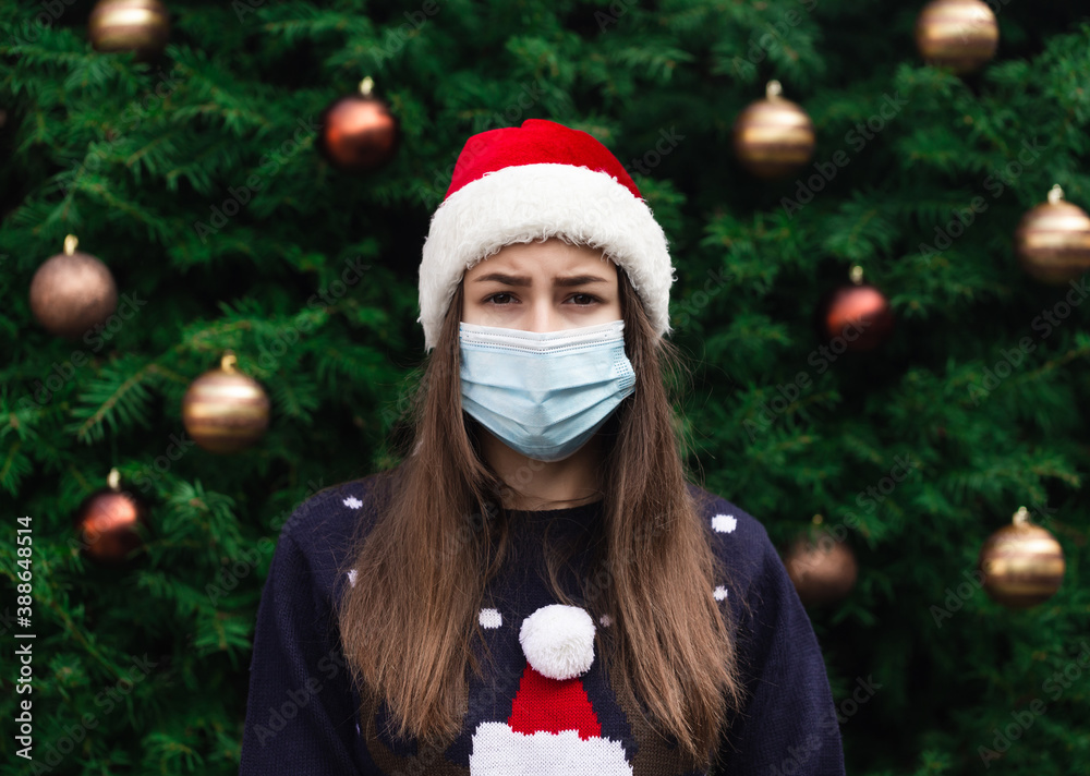 Close up Portrait of woman wearing a santa claus hat and medical mask with emotion. Against the background of a Christmas tree. Coronavirus pandemic Close up Portrait of woman wearing a santa claus hat and medical mask with emotion. Against the background of a Christmas tree. Coronavirus pandemic