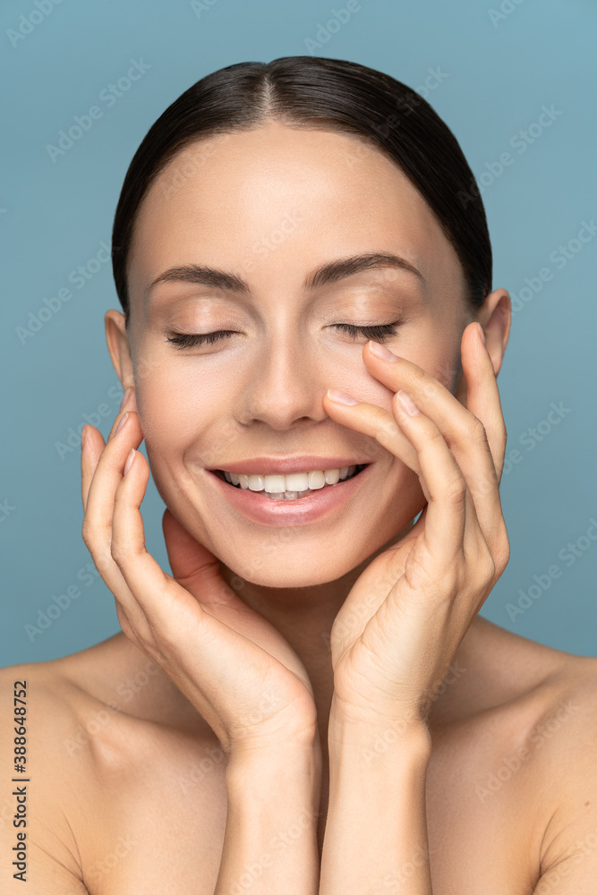 Young woman with natural makeup, combed hair, touching her well-groomed pure skin on face, closed eyes, smiling, standing over studio blue background. Beauty, facelift, anti-aging cosmetic procedure.