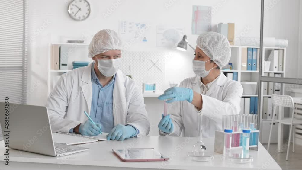 Slow mo medium of two doctors wearing medical masks, gloves, caps sitting by desk in lab, female senior Asian worker holding test tube talking to male Caucasian colleague taking notes in journal Slow mo medium of two doctors wearing medical masks, gloves, caps sitting by desk in lab, female senior Asian worker holding test tube talking to male Caucasian colleague taking notes in journal
