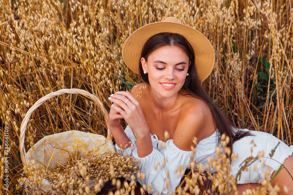 Young beautiful woman with straw hat on the head laying at golden oat field holding basket with ears of oats.
