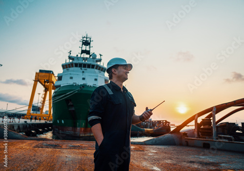 Marine Deck Officer or Chief mate on deck of offshore vessel or ship , wearing PPE personal protective equipment - helmet, coverall. He holds VHF walkie-talkie radio in hands.