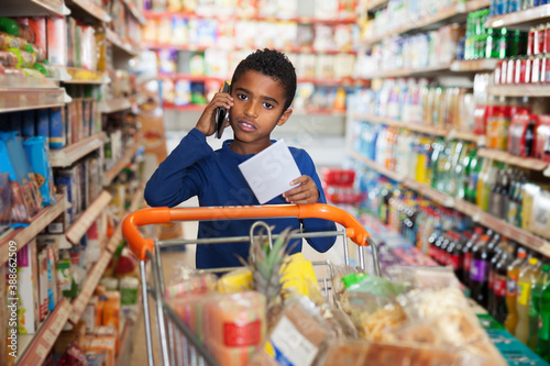 African American preteen boy choosing food goods with shopping list and talking by phone in store
