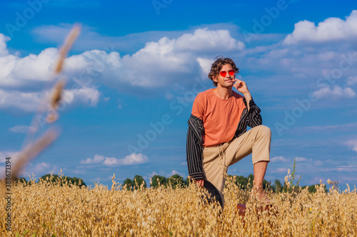 Tall handsome man with black hat and pink sunglasses standing with one leg on a brown vintage leather suitcase at golden oat field. © Smile