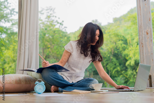 Asian woman reading order on laptop and writing a document in living room and background blur beautiful garden at home. (lens blur effect)