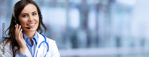 Portrait of happy smiling young female doctor in photo headset, over blurred office background, with blank copy space area for some slogan or text. Medical call center concept picture.