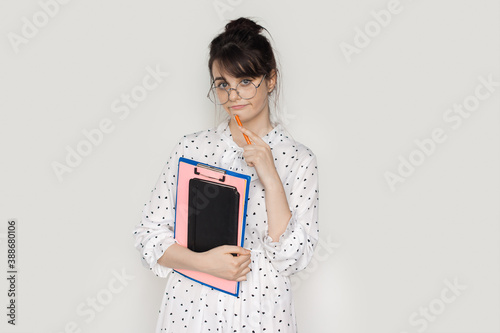 Brunette woman with glasses is holding some folders and posing on a white studio wall