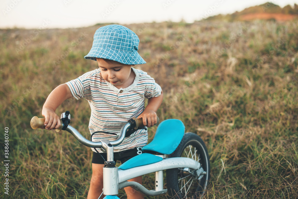 Obraz premium Caucasian small boy walking with a bike on a field wearing a blue hat in a summer day