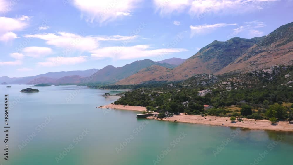 The shoreline of lake Skadar,Montenegro,with its clear waters,high mountain ranges around,forests and houses on the beach,white clouds and sunny skies above,small island in the distance,aerial view. The shoreline of lake Skadar,Montenegro,with its clear waters,high mountain ranges around,forests and houses on the beach,white clouds and sunny skies above,small island in the distance,aerial view.