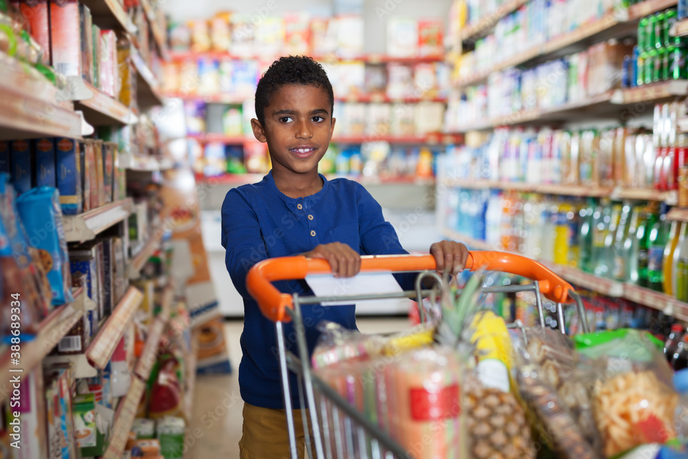 Positive preteen African boy carrying full grocery cart after shopping in grocery store