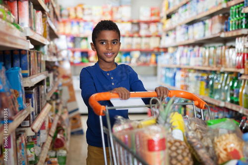 Positive preteen African boy carrying full grocery cart after shopping in grocery store