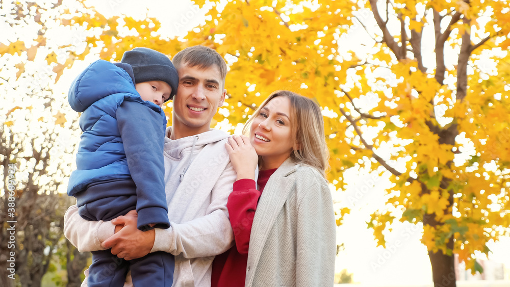 Lovely family of mother father and funny little boy hug posing for camera in picturesque autumn city park on nice warm day low angle shot Lovely family of mother father and funny little boy hug posing for camera in picturesque autumn city park on nice warm day low angle shot