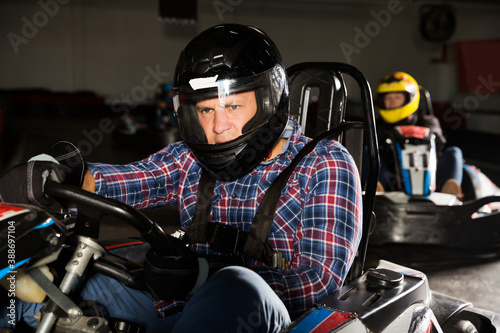 active adult man in helmet and other people driving car for karting in sport club indoor