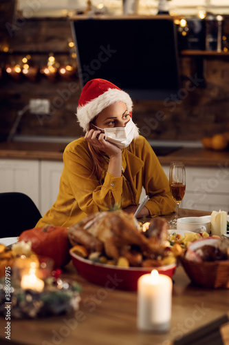 Pensive woman with face mask sitting alone at dining table on Christmas. © Drazen