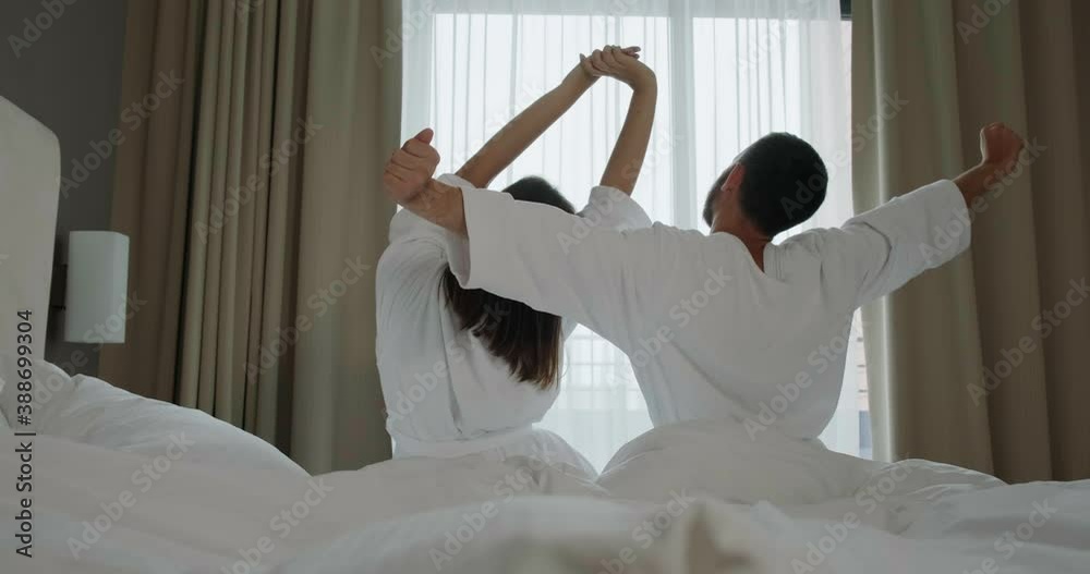 Cute couple stretches and kisses sitting on large comfortable bed in hotel room in morning at honey moon backside view Cute couple stretches and kisses sitting on large comfortable bed in hotel room in morning at honey moon backside view