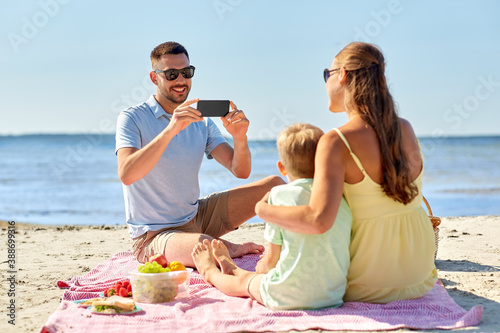 family, leisure and people concept - happy father with smartphone photographing mother and son on summer beach