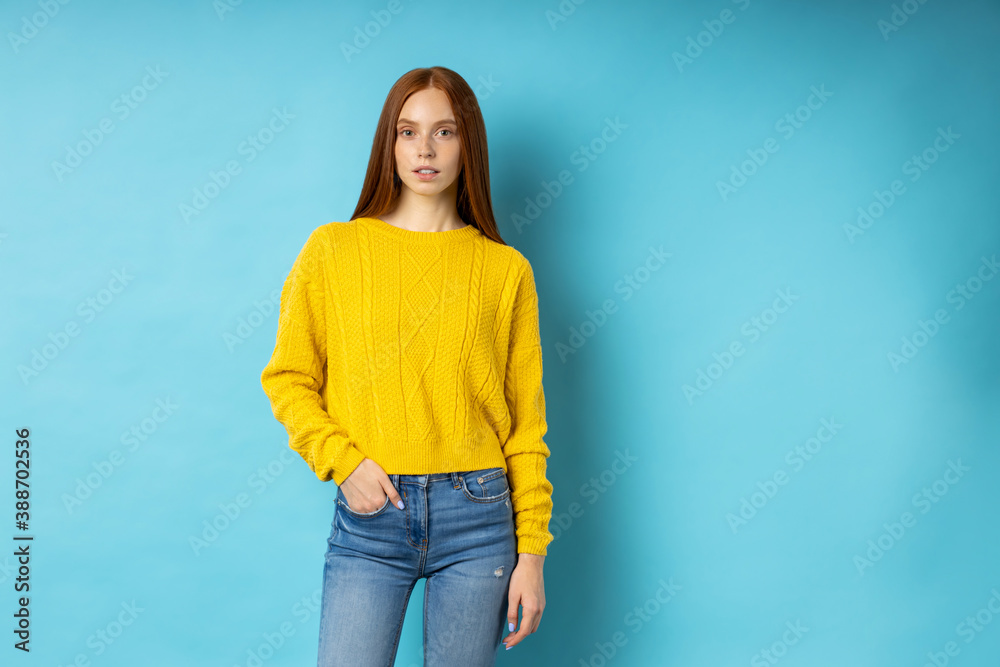 Portrait of redhead attractive slim caucasian serious calm young lady wearing yellow sweater, jeans keeping hand in pocket posing on blue background with copy space. Portrait of redhead attractive slim caucasian serious calm young lady wearing yellow sweater, jeans keeping hand in pocket posing on blue background with copy space.