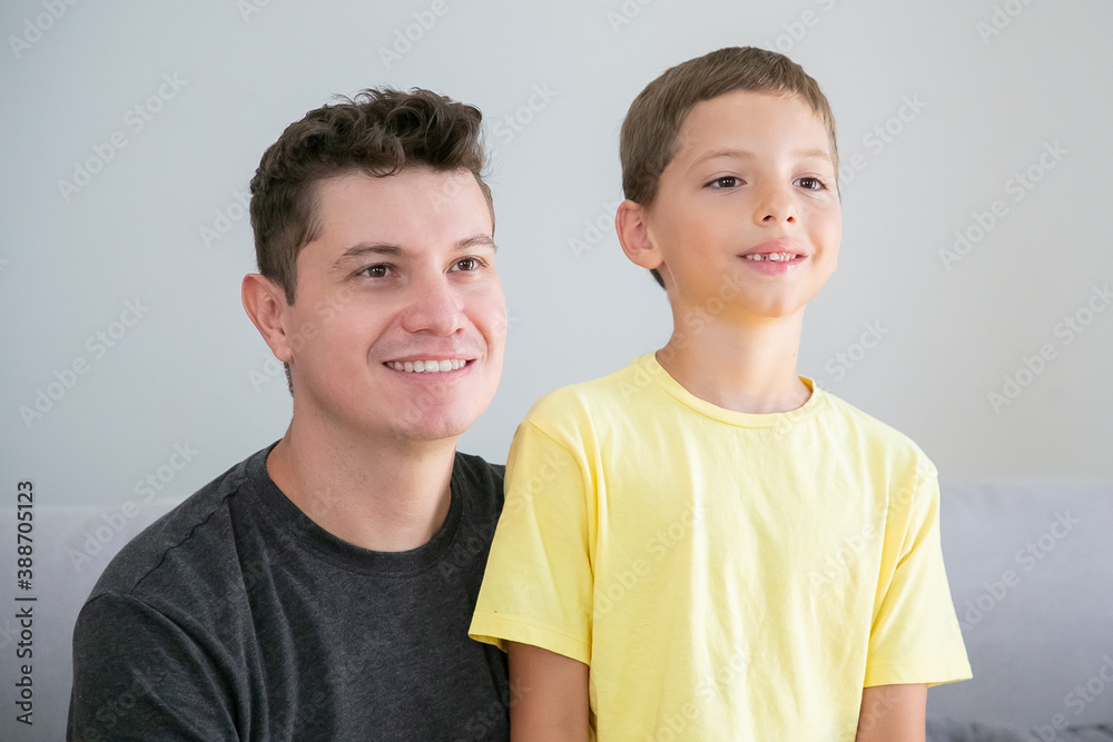 Happy boy and his dad sitting together indoors and looking away. Medium shot. Family and parenthood concept Happy boy and his dad sitting together indoors and looking away. Medium shot. Family and parenthood concept