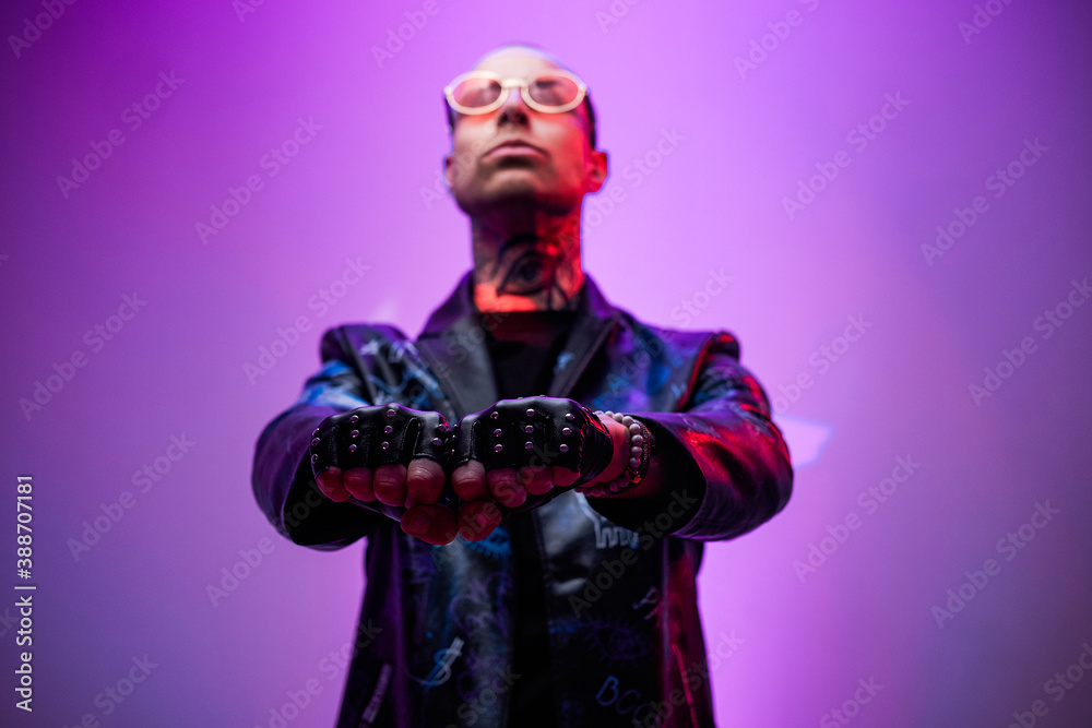 Serious and styled rocker in black leather jacket with glasses pulling his hands forward and poses in abstract background.