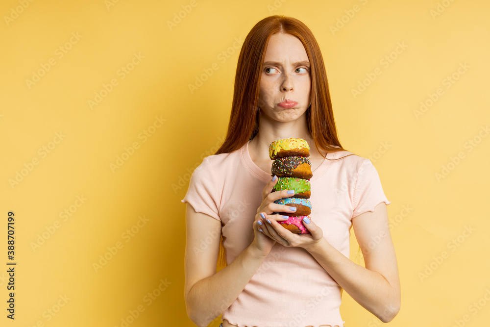 Waist up shot of serious caucasian woman with long red hair, in t shirt holding colorful glazed donuts, looking strictly at camera over yellow background. Unhealthy food, dieting, concept. Waist up shot of serious caucasian woman with long red hair, in t shirt holding colorful glazed donuts, looking strictly at camera over yellow background. Unhealthy food, dieting, concept.