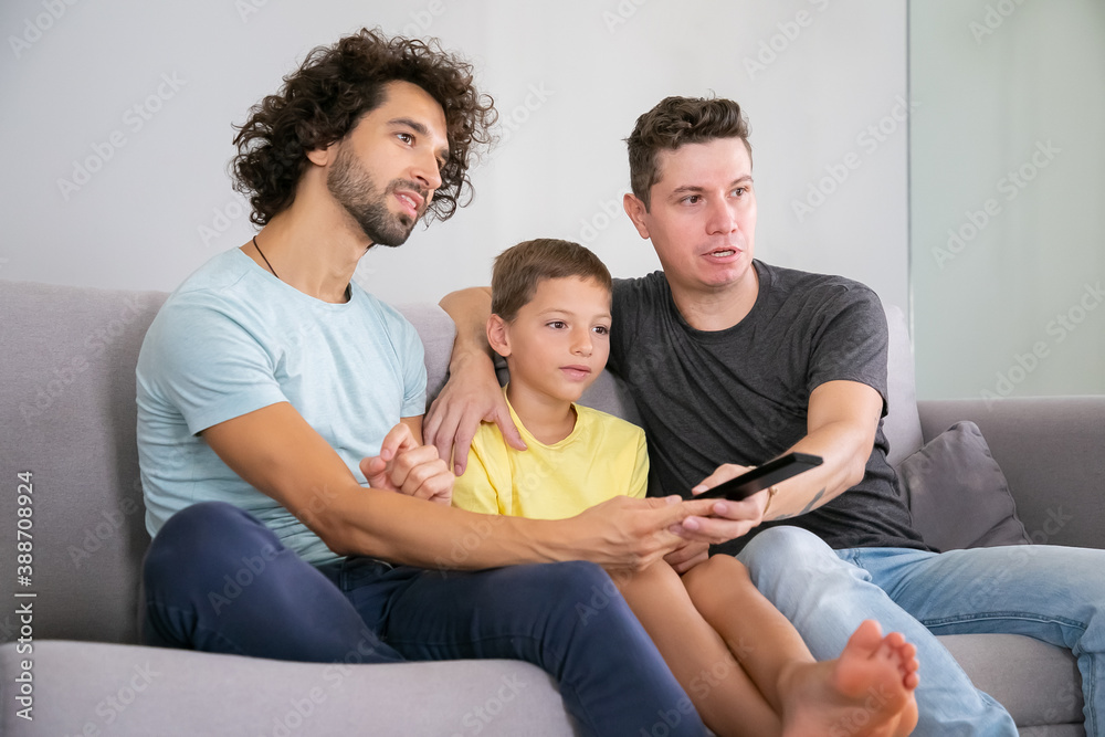 Excited homosexual dads and son watching TV show at home, sitting on couch in living room, hugging, using remote control, looking away. Family and home entertainment concept Excited homosexual dads and son watching TV show at home, sitting on couch in living room, hugging, using remote control, looking away. Family and home entertainment concept