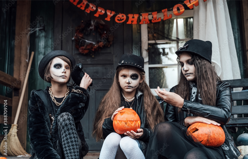 Scary girls, sisters, friends celebrating halloween.Treak or treat game.Pumpkins.Sitting on porch steps.Bags with sweets in hands.Terrifying face skull makeup.Witch stylish costumes. Children's party