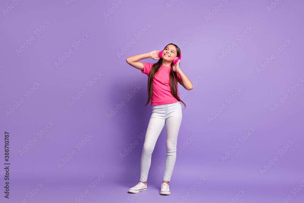 Full length body size photo of dreamy little girl smiling listening to favourite songs with headphones wearing t-shirt jeans isolated on purple color background Full length body size photo of dreamy little girl smiling listening to favourite songs with headphones wearing t-shirt jeans isolated on purple color background