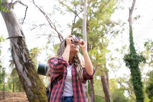 Blonde young lady shooting landshate and walking in forest. Female traveler exploring nature, holding camera, taking photo and carrying big backpack. Tourism, adventure and summer vacation concept