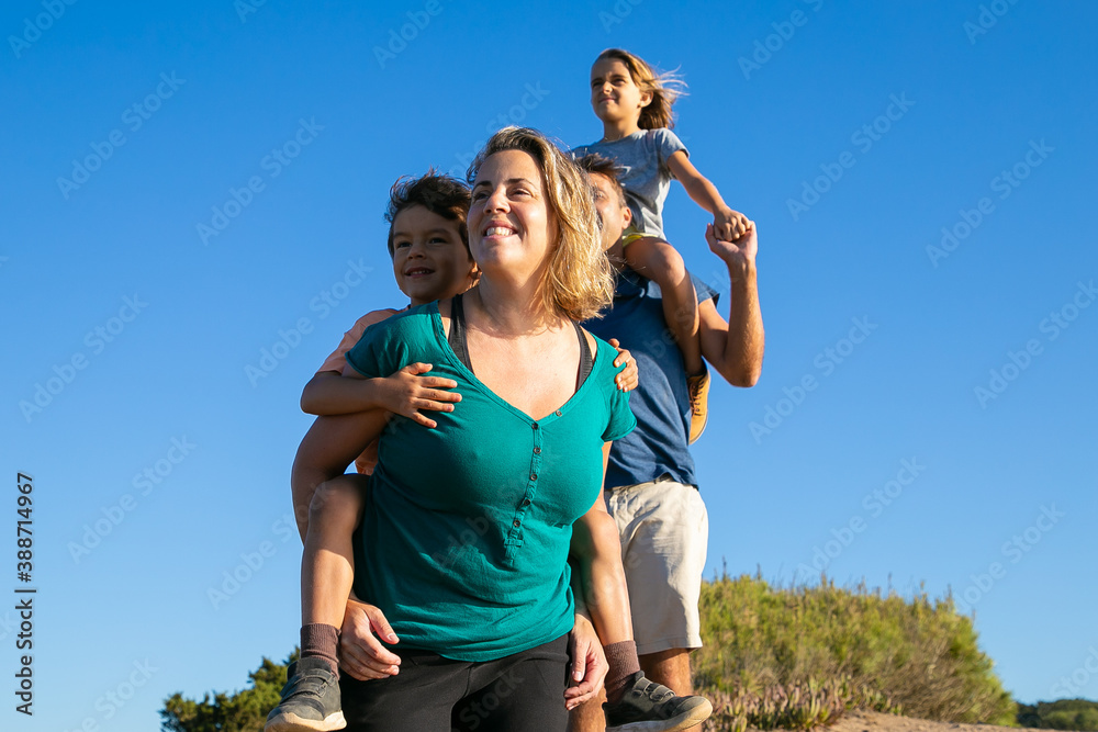 Obraz premium Happy family enjoying hiking in countryside. Two kids riding on parents back and neck. Low angle. Nature and recreation concept