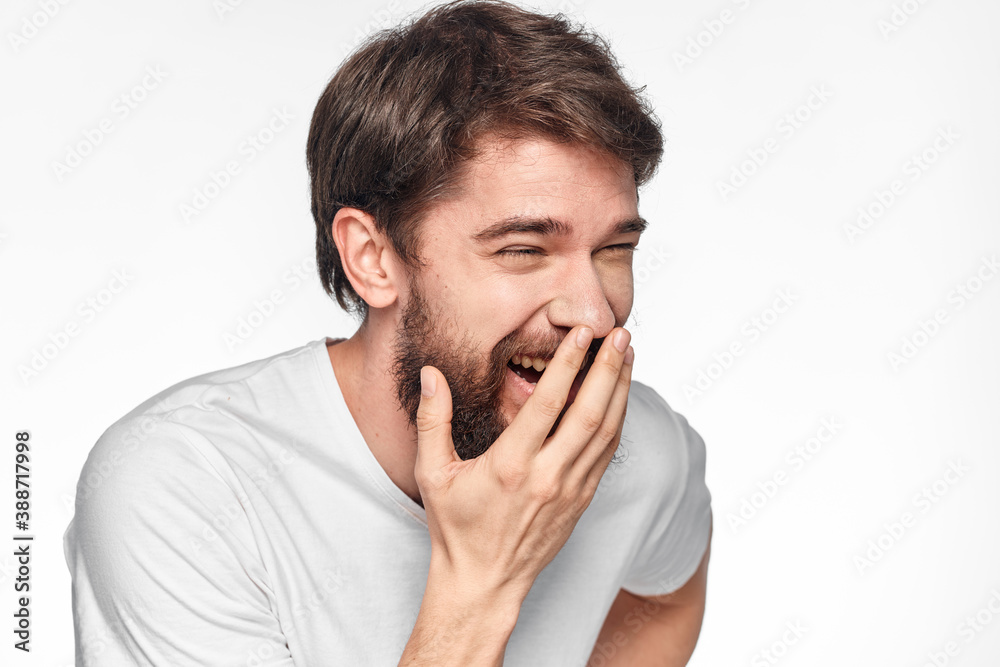 cheerful bearded man in a white t-shirt emotions gestures with his hands light background studio