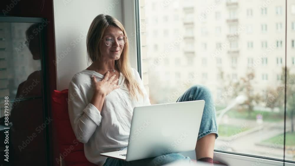 A young woman with glasses taking a video call with her laptop, and drinks coffee from a mug while sits on the windowsill with a city view. A remote employee working from home. A young woman with glasses taking a video call with her laptop, and drinks coffee from a mug while sits on the windowsill with a city view. A remote employee working from home.