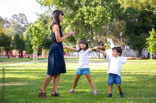 Two happy cute kids and their mom playing active games outdoors, doing exercises on grass in park. Family outdoor activity and leisure concept