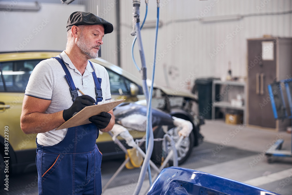 Bearded auto mechanic writing on clipboard at vehicle repair shop Bearded auto mechanic writing on clipboard at vehicle repair shop