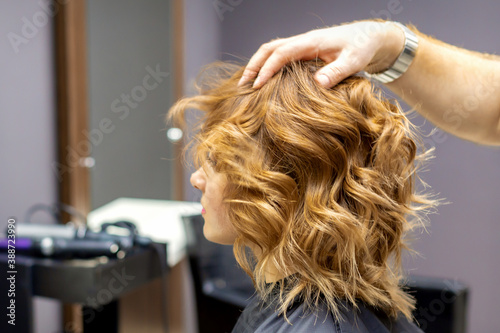 Side view of male hairdresser checks curly hairstyle of young caucasian woman in hair salon. Soft focus