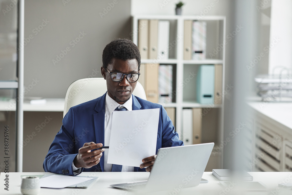 African young businessman in eyeglasses sitting at his workplace in front of laptop and reading a contract