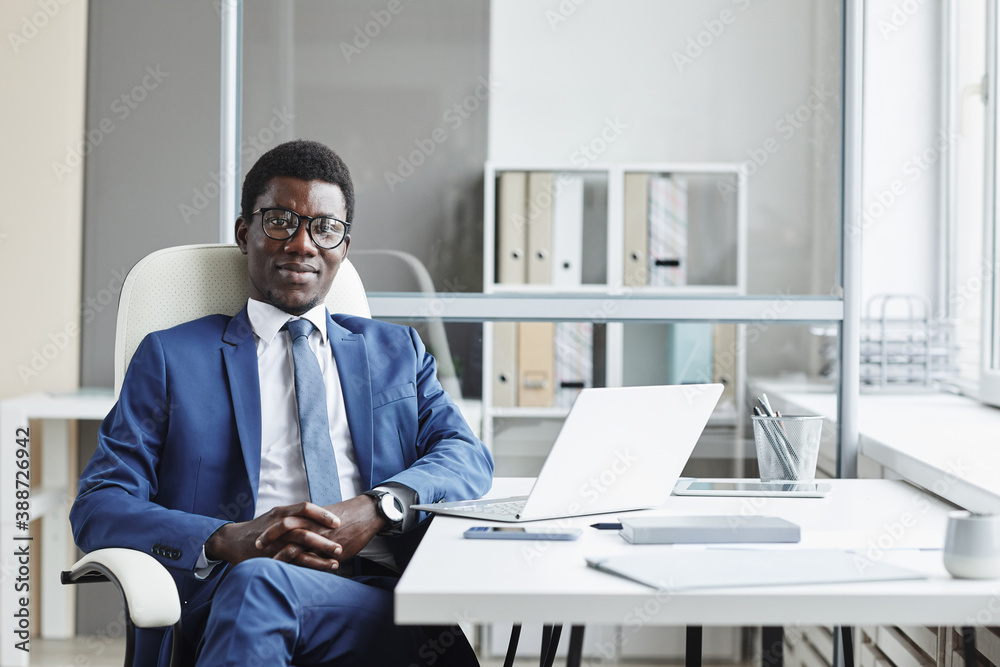 Fototapeta premium Portrait of African businessman in suit sitting on chair at his workplace and looking at camera at office