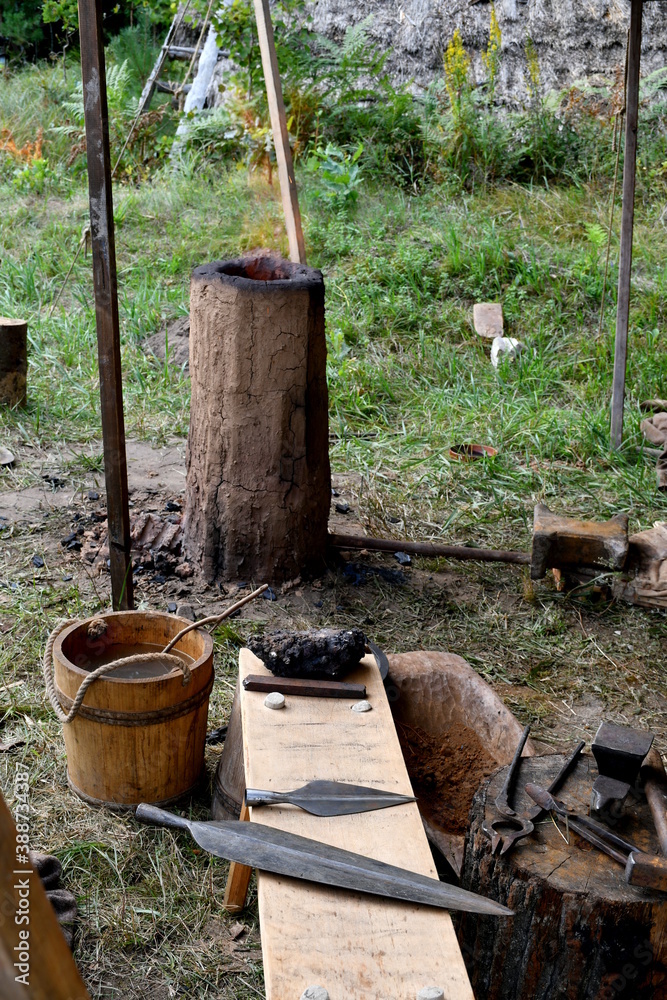 A close up on a temporary blacksmith workshop with a clay chimney used for melting steel, bucket of water, and copper weapon elements on a wooden bench seen during a history fair in Poland
