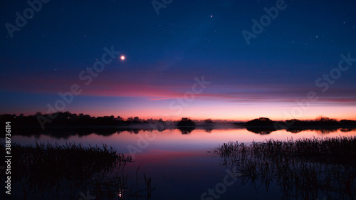 Сolorful sunset on the shore of a foggy lake, river with grass and bushes trees and houses on the shore, light in the windows, a starry night with a moon in the sky, clouds reflected in the water