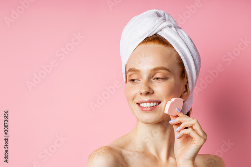 young joyful woman with white towel on head, fresh pure skin keeping cosmetic sponge near face, smiling looking aside on pink background with copy space for text, advertising. Beauty products.